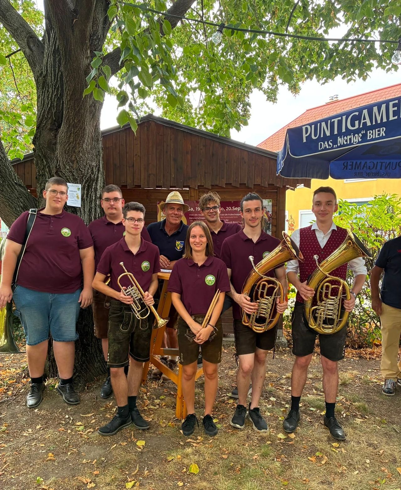 Eine Gruppe von Musikern in burgunderfarbenen Hemden und Shorts posiert für ein Foto vor einem Baum und einem Gebäude mit einem blauen Vordach. Sie halten Blechblasinstrumente, und eine Person steht hinter einem Tisch.
