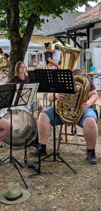 Eine Gruppe von Musikern spielt im Freien mit einem Tuba, Trommeln und Notenständern. Eine Frau und ein Mann spielen, während ein Kind im Hintergrund ist.