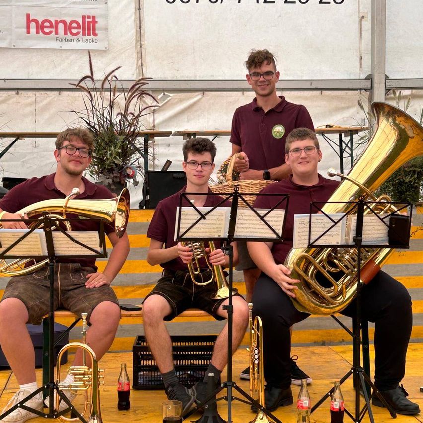 Vier junge Männer, alle in burgunderroten Hemden und Shorts gekleidet, sitzen auf einer Bühne und halten jeweils ein Blechblasinstrument. Hinter ihnen steht ein Mann mit einem Korb. Ein Banner mit dem Text 'henelit' ist im Hintergrund zu sehen.