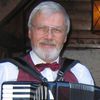 Ein älterer Mann mit Brille und roter Fliege lächelt, während er ein Akkordeon spielt, in einer hölzernen Umgebung.
