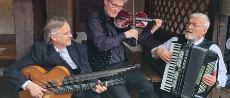 Drei Musiker sitzen im Freien, einer spielt Geige, ein anderer Gitarre und der dritte Akkordeon. Sie sind in einer hölzernen Kabine.
