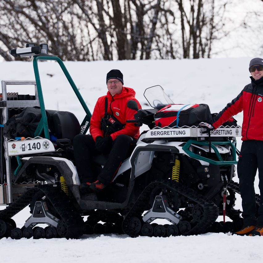 Zwei Personen stehen neben einem Schneemobil, einer sitzt, beide sind in roten und schwarzen Jacken gekleidet. Das Schneemobil hat ein Nummernschild mit der Nummer 140 und angebrachte Taschen. Sie befinden sich in einem verschneiten Bereich mit Bäumen im Hintergrund.