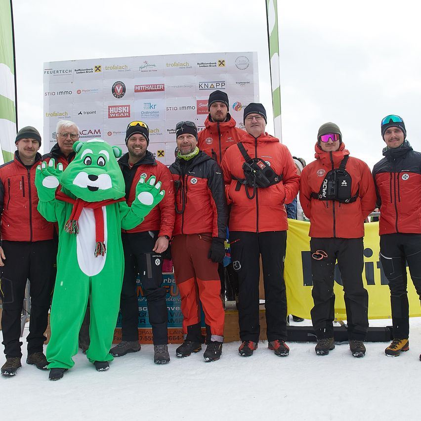 Eine Gruppe von Menschen in roten Jacken und Schneestiefeln steht vor einem Banner mit Logos, einer trägt ein grünes Maskottchenkostüm.