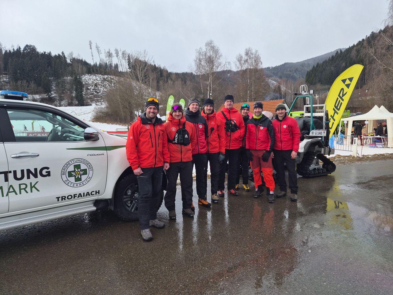Eine Gruppe von Menschen in roten Jacken steht vor einem Auto. Sie stehen auf nassem Pflaster. Im Hintergrund befindet sich ein Berg mit Schnee, Bäumen und einem gelben Banner.