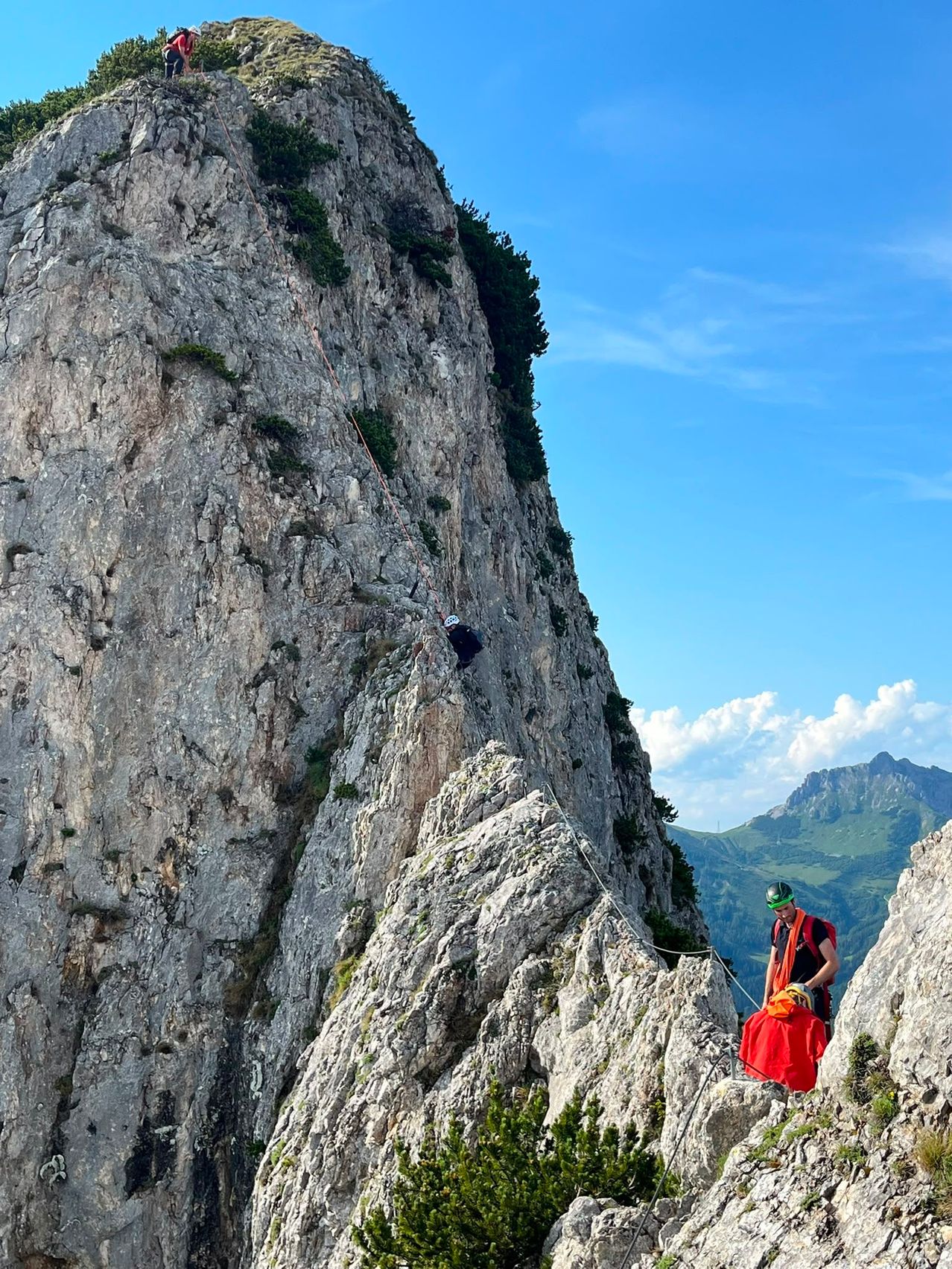 Zwei Personen klettern an einem steilen Berg, gesichert durch Seile. Ein Kletterer ist höher oben am Berg, während der andere tiefer ist und eine rote Tasche hält.