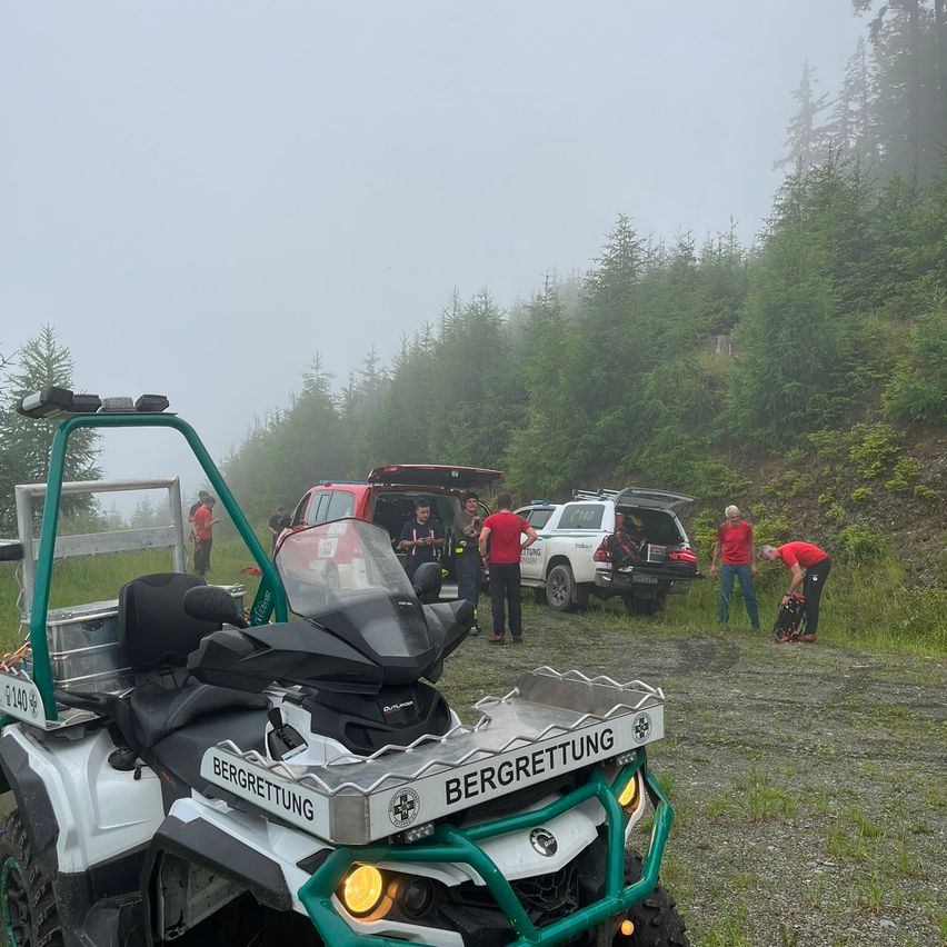 Eine Gruppe von Personen in roten Shirts führt einen Einsatz mit einem Rettungsfahrzeug auf einer Schotterstraße im Wald durch.