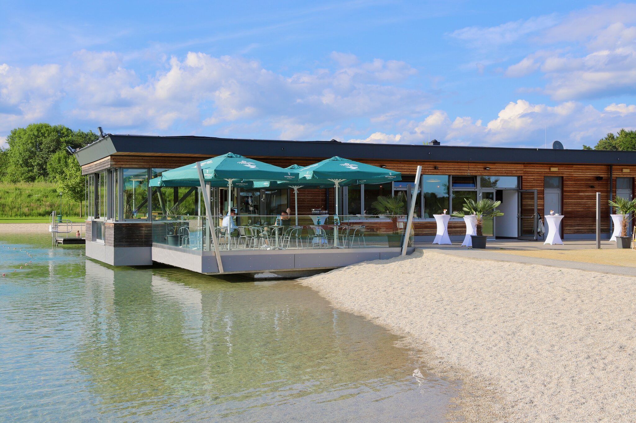 Ein Restaurant am Wasser mit grünen Sonnenschirmen, Tischen, Stühlen und Topfpflanzen. Das Gebäude hat Glaswände und ein schwarzes Dach.