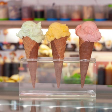 Drei Eisbecher in weiß, gelb und pink sind auf einem Glasständer vor einem Geschäft ausgestellt.