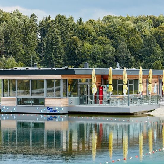 Ein modernes schwimmendes Gebäude mit dem Namen See Werk an der Seite, ausgestattet mit Glasfenstern, einer Terrasse mit Tischen und Stühlen sowie gelben Sonnenschirmen. Das Gebäude ist von Bäumen und einem ruhigen See umgeben.