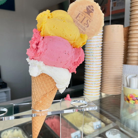 Ein Eisbecher mit drei Schichten Eis. Die untere Schicht ist weiß, die mittlere rosa und die oberste gelb.