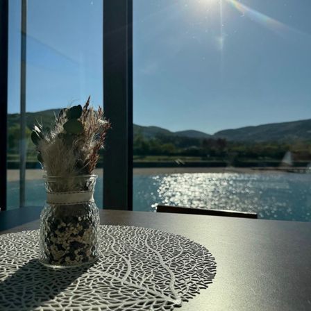 Ein klarer Tag mit Sonnenstrahlen, die durch ein Glasfenster scheinen und einen Blick auf einen See und Berge zeigen. Ein Glas mit getrockneten Pflanzen steht auf einem Tisch und reflektiert Sonnenlicht.