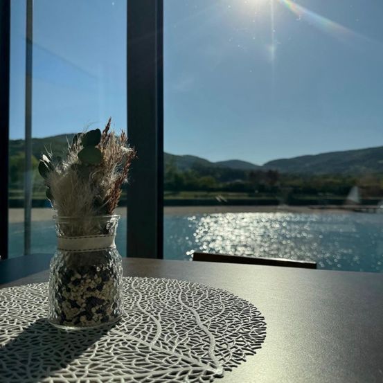 Ein klarer Tag mit Sonnenstrahlen, die durch ein Glasfenster scheinen und einen Blick auf einen See und Berge zeigen. Ein Glas mit getrockneten Pflanzen steht auf einem Tisch und reflektiert Sonnenlicht.