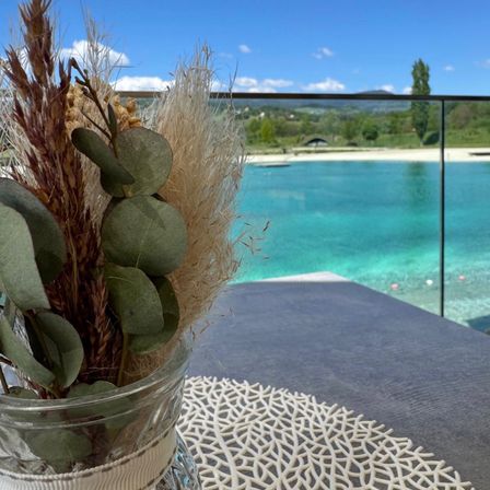 Ein Glasvase mit getrockneten Pflanzen steht auf einem Tisch mit einer weißen Spitzendeckchen und bietet einen Blick auf einen Pool. Der Pool hat klares blaues Wasser und einen Blick auf eine grüne Landschaft.