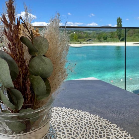 Ein Glasvase mit getrockneten Pflanzen steht auf einem Tisch mit einer weißen Spitzendeckchen und bietet einen Blick auf einen Pool. Der Pool hat klares blaues Wasser und einen Blick auf eine grüne Landschaft.