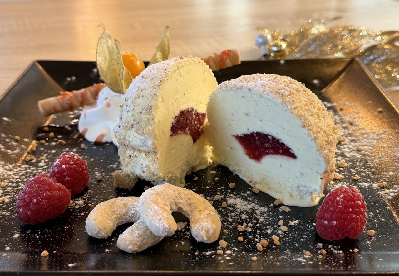 Ein Dessert auf einem schwarzen Teller besteht aus einem geschnittenen Käsekuchen mit Himbeerfüllung, bestreut mit Puderzucker, umgeben von frischen Himbeeren und Keksen.