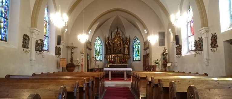 Ein traditionelles Kircheninneres mit Buntglasfenstern, hölzernen Kirchenbänken, einem roten Teppich zum Altar und verschiedenen religiösen Artefakten und Statuen.