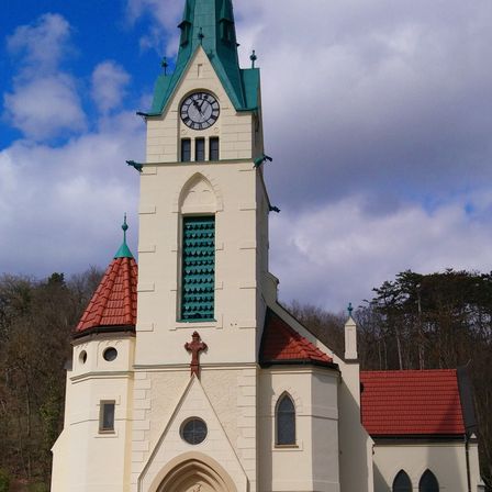 Ein hoher Kirchturm mit einem grünen Spitzturm und einer Uhr, umgeben von Bäumen unter einem bewölkten Himmel.