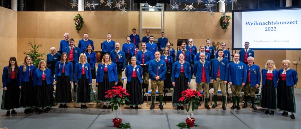Ein Chor in traditioneller Kleidung steht auf der Bühne, mit festlichen Dekorationen und einer Projektionsleinwand mit 'Weihn' dahinter.