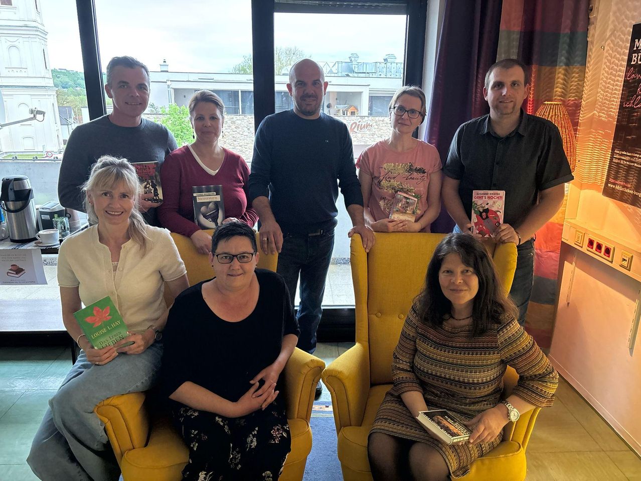 Sieben Personen sitzen in gelben Sesseln und posieren für ein Foto. Zwei Personen stehen hinter ihnen. Eine Frau auf der linken Seite hält ein Buch, und ein Mann auf der rechten Seite hält ein Buch und lächelt.