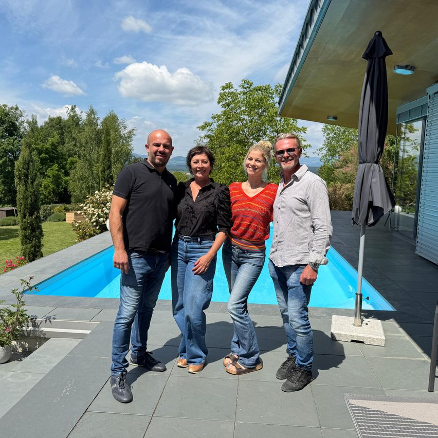 Vier Personen stehen vor einem modernen Haus mit Pool und lächeln für ein Foto. Hinter ihnen befindet sich eine Rasenfläche mit Bäumen und einem geschlossenen Schirm.