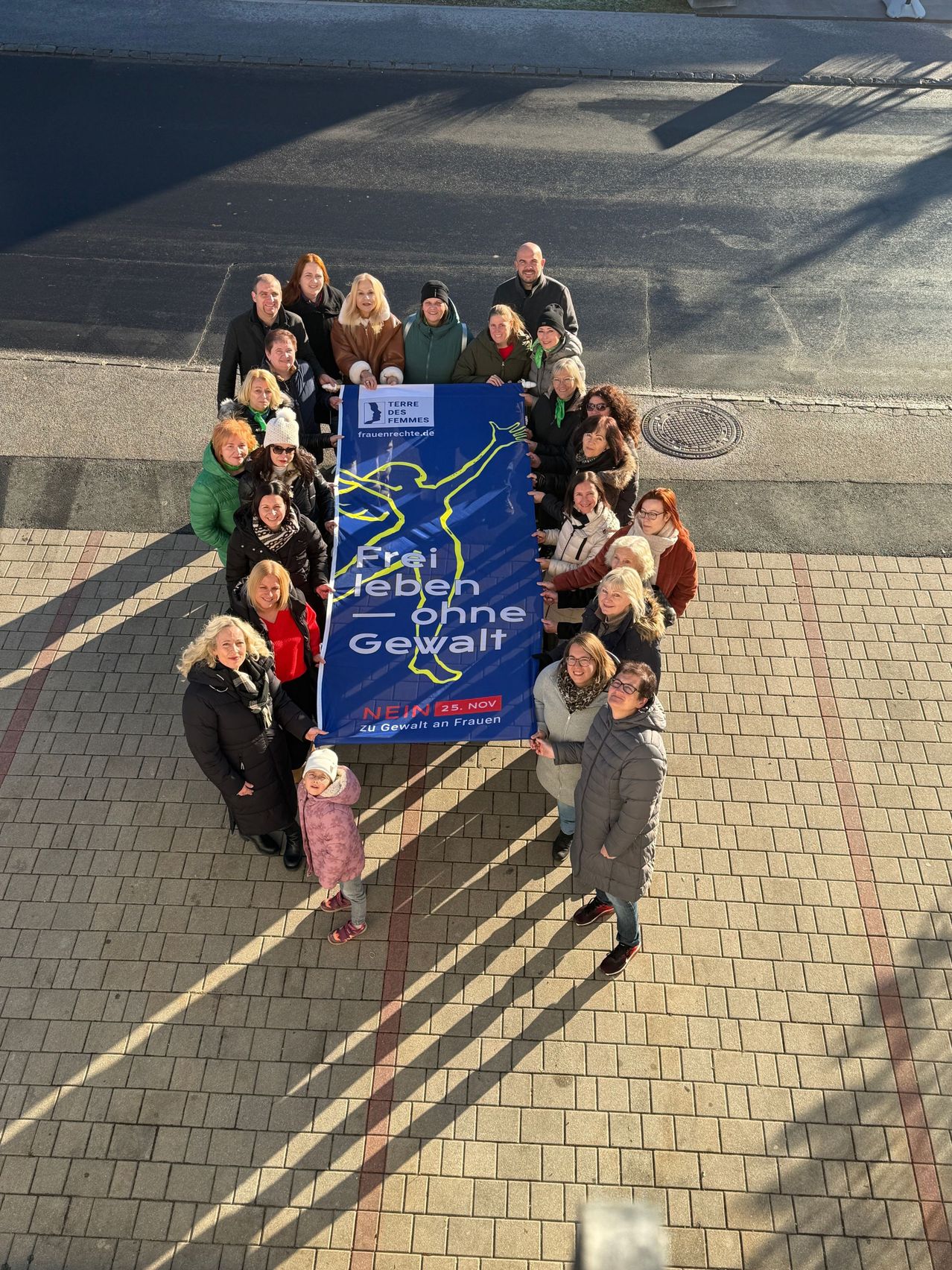 Eine Gruppe von Menschen steht im Kreis und hält ein blaues Banner mit weißen Buchstaben 'Frei leben ohne Gewalt.' Sie posieren für ein Foto auf einem Ziegelweg.