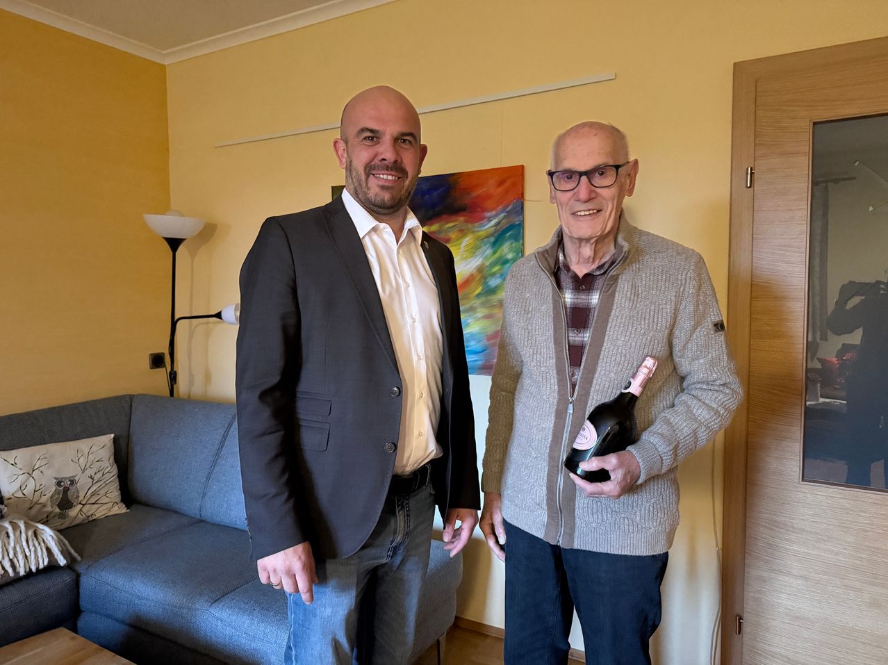 Zwei Männer stehen in einem gelb gestrichenen Raum. Einer hält eine Flasche Wein und lächelt, während der andere einen Anzug trägt. Ein Sofa, eine Lampe und ein Gemälde im Hintergrund.