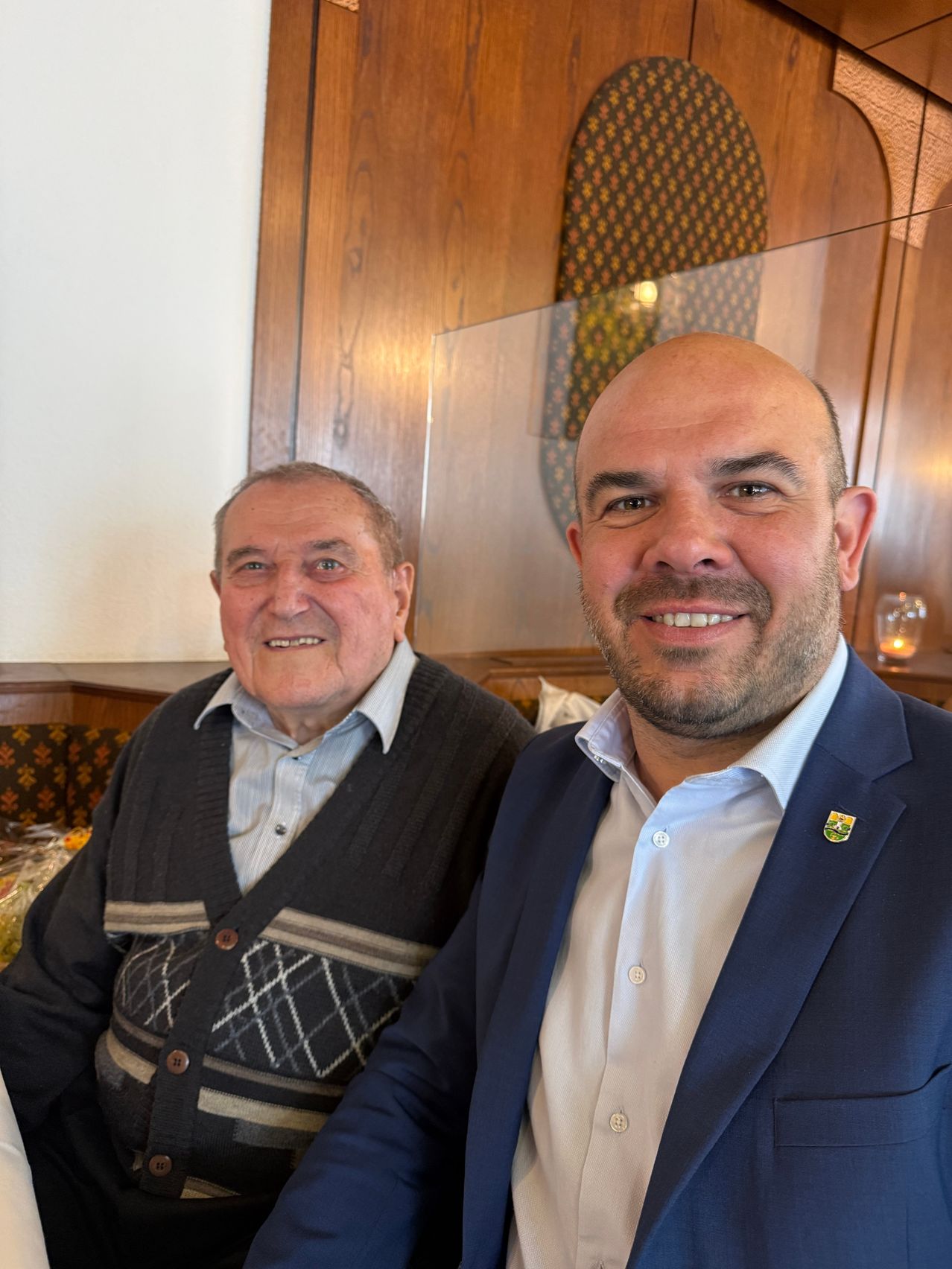 Zwei Männer sitzen in einer Kabine, lächelnd, einer trägt einen Pullover, der andere einen Anzug mit Abzeichen. Hinter ihnen eine Holzwand und eine Glasbarriere.