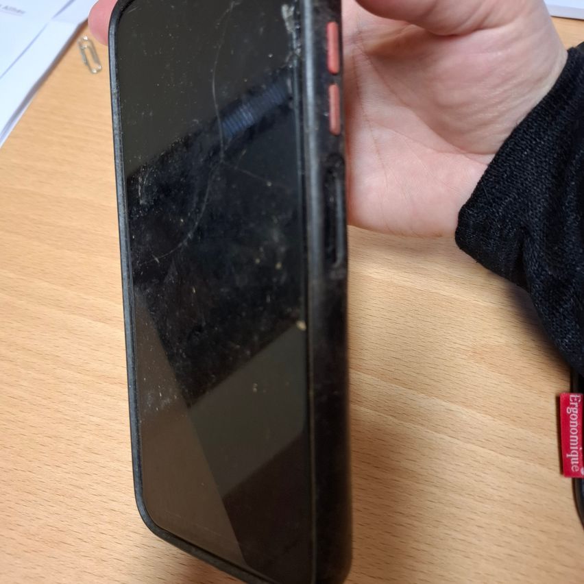 Eine Person hält ein beschädigtes schwarzes Telefon mit einem gebrochenen Bildschirm auf einem Holztisch.