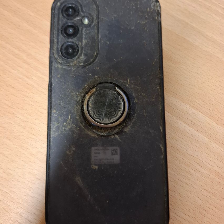 Rückseite eines Mobiltelefons mit beschädigter Kamera und Lautsprecher. Es hat einen runden Knopf und einen kleinen quadratischen Barcode. Das Gerät liegt auf einer Holzoberfläche.