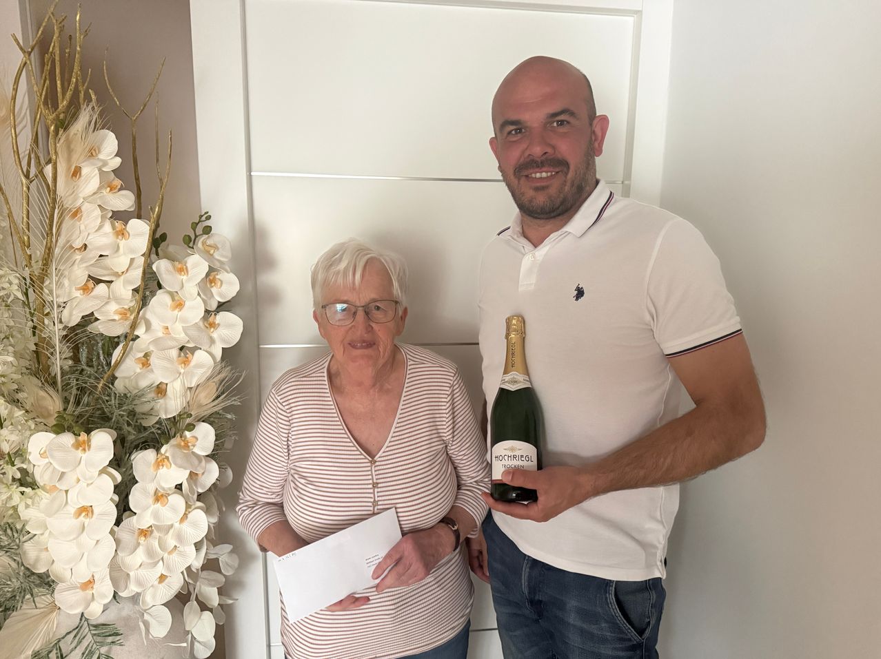 Ein Mann und eine Frau stehen lächelnd nebeneinander, der Mann hält eine Flasche Champagner. Die Frau hält einen weißen Umschlag. Ein weißer Orchideenstrauß steht hinter ihnen.