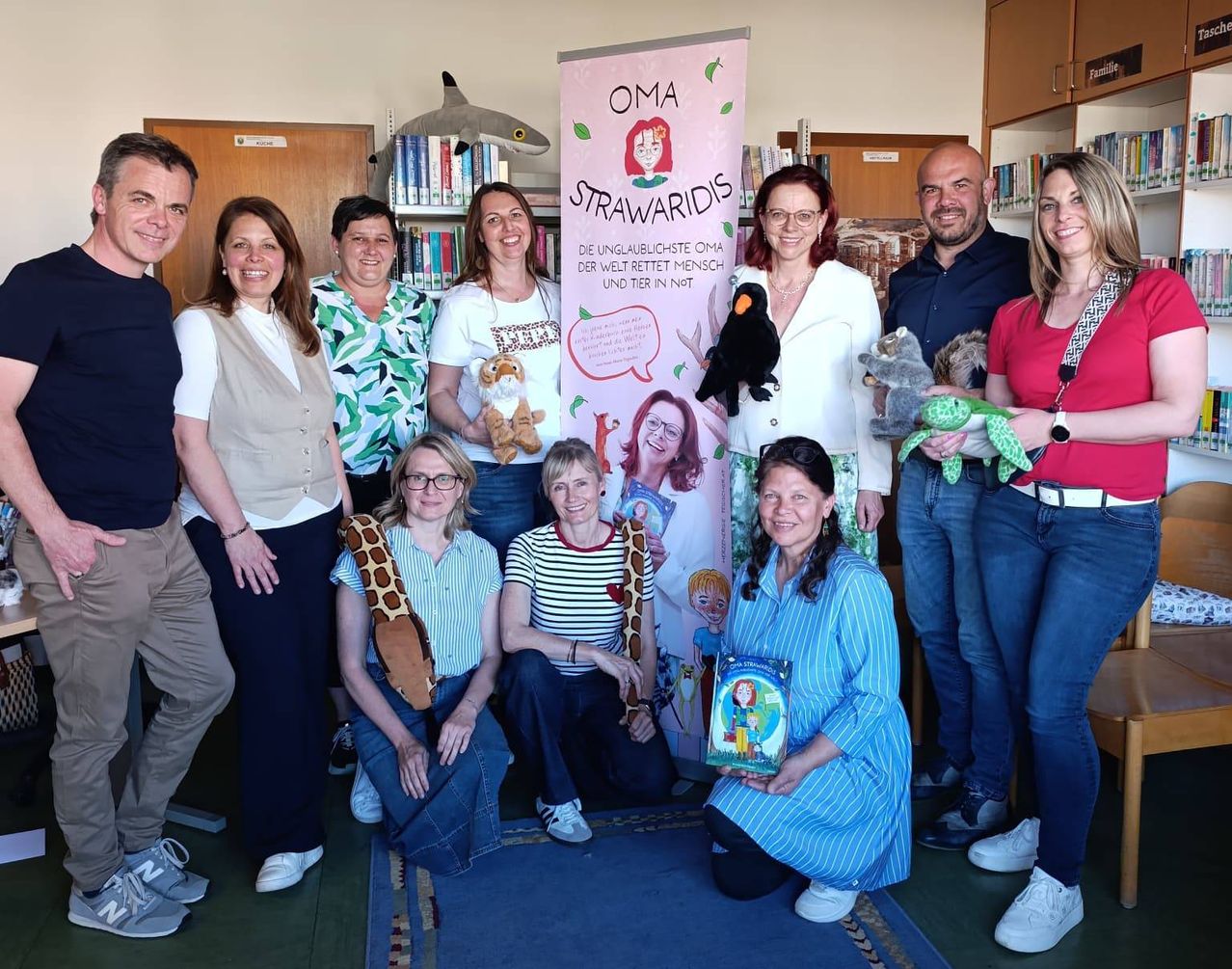 Eine Gruppe Erwachsener und Kinder posiert mit Plüschtieren und einem Buch mit dem Titel 'OMA STRAWARIDIS' in einer Bibliothek.