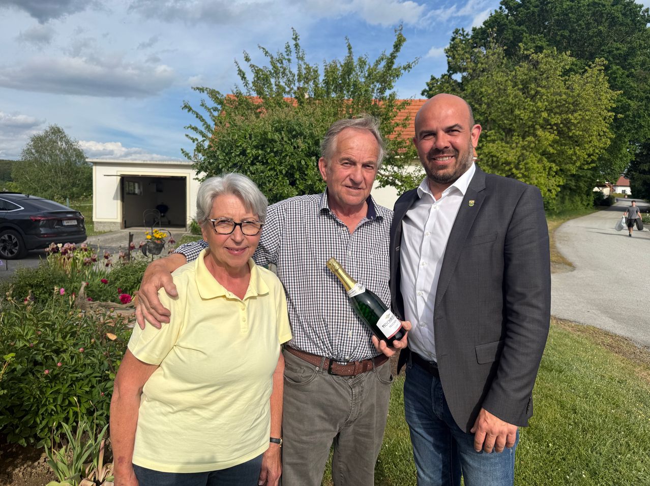 Drei Personen stehen in einem Garten. Ein Mann hält eine Flasche Champagner. Eine Frau und ein Mann lächeln.