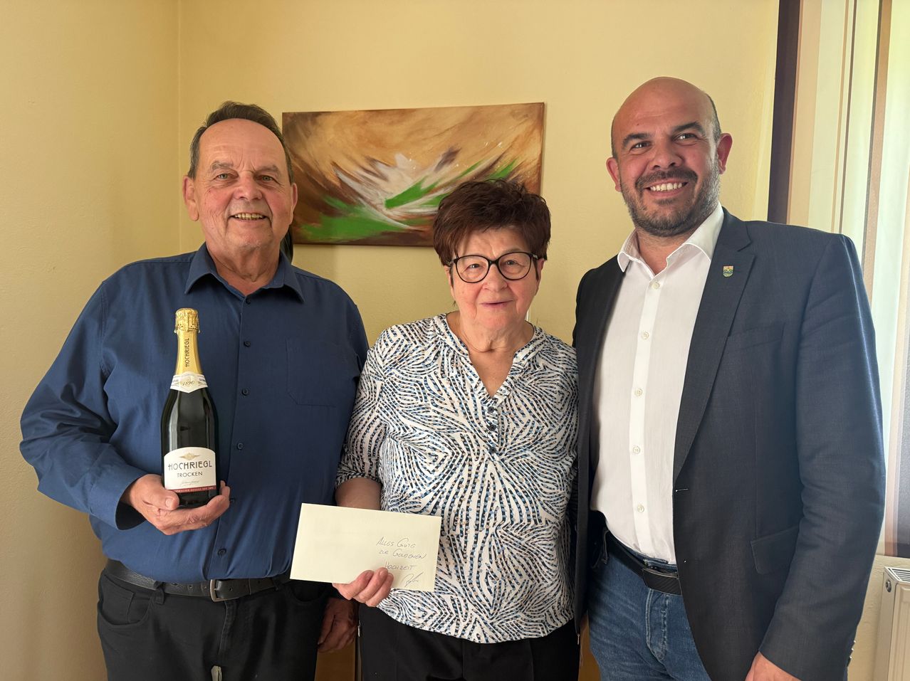 Drei Personen posieren für ein Foto. Ein Mann hält eine Flasche Wein und ein Zertifikat. Eine Frau hält das Zertifikat, und ein Mann steht neben ihr. Sie alle lächeln. Ein Gemälde ist an der Wand hinter ihnen.