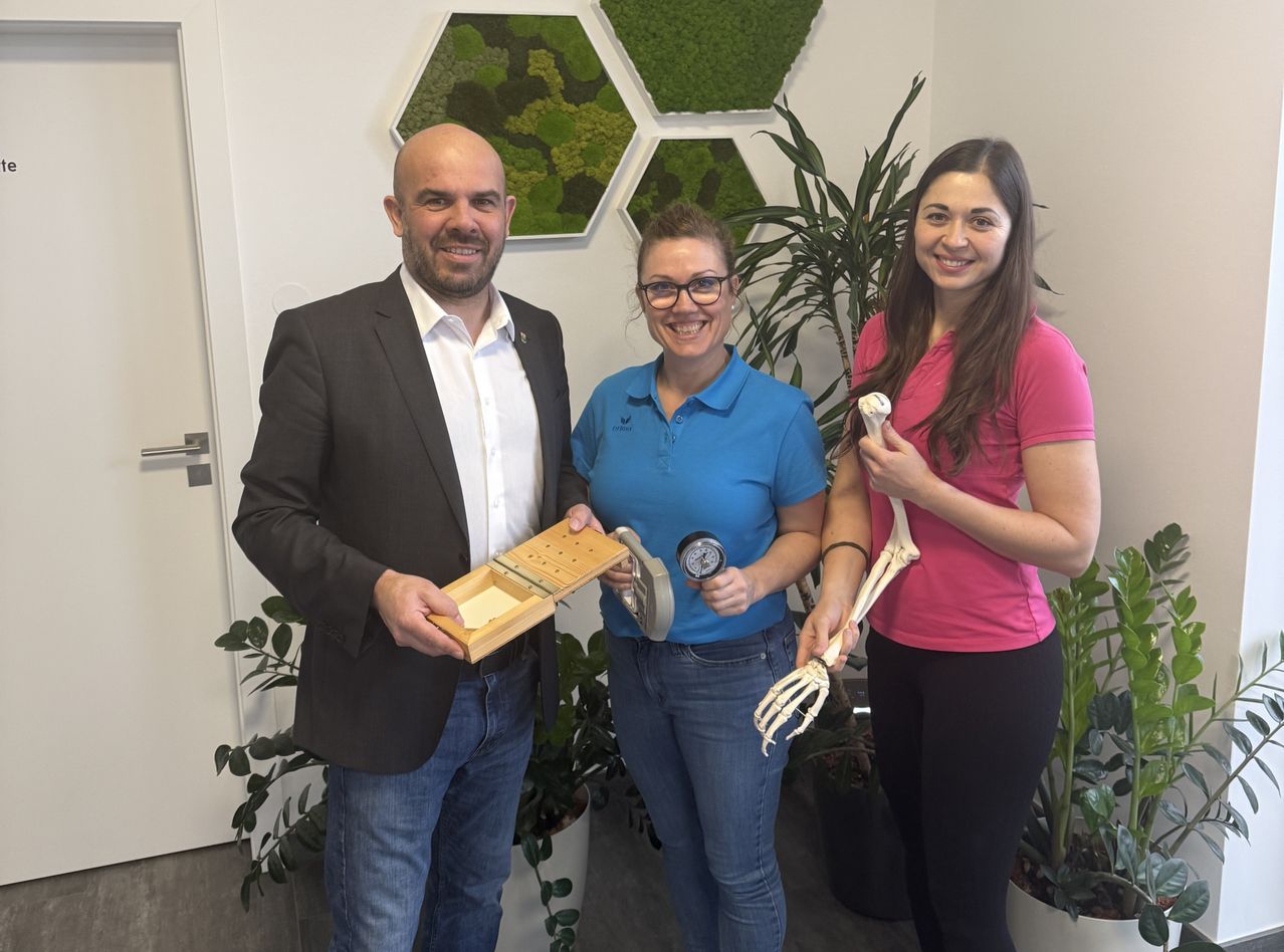 Drei Personen stehen in einem Büro und lächeln. Ein Mann hält eine Holzkiste, eine Frau ein Stethoskop und eine weitere Frau ein Skelett. Sie sind von Pflanzen umgeben.