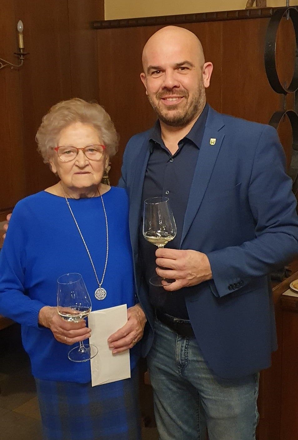 Eine ältere Frau und ein Mann lächeln und posieren für ein Foto, während sie Weingläser halten. Sie stehen eng beieinander, wahrscheinlich bei einer Veranstaltung. Die Frau trägt eine Brille und eine Halskette. Der Mann hat einen Blazer mit einem Logo auf der linken Brust. Hinter ihnen ist eine Holzwand.