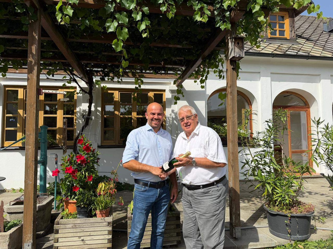 Zwei Männer stehen unter einer Pergola, einer hält eine Flasche Wein und lächelt vor einem weißen Gebäude mit Bogenfenstern und viel Grün.