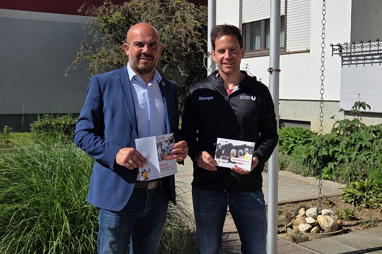 Zwei Männer stehen lächelnd draußen. Einer hält einen Prospekt mit dem Titel Unser Angebot und der andere einen mit dem Titel Vereinsservice. Sie stehen vor einem Gebäude.