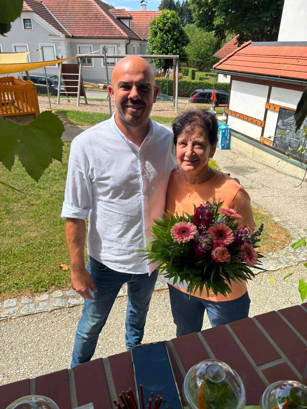 Ein lächelnder Mann und eine Frau stehen draußen, der Mann hält einen Blumenstrauß. Dahinter befindet sich ein Spielplatz mit einer Rutsche und einem geparkten Auto. Ein Haus mit einer Tafel und einer Rebe befindet sich rechts.