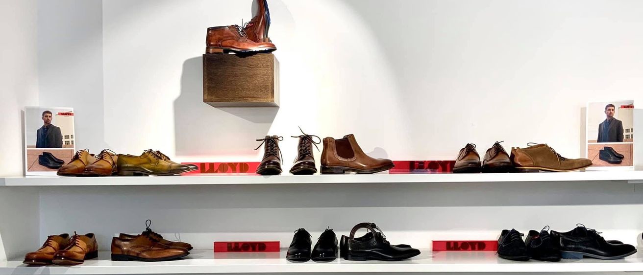 Eine Ausstellung verschiedener brauner und schwarzer Lederschuhe, einschließlich Stiefel und Geschäftsschuhe, auf weißen Regalen angeordnet. Ein brauner Schuh ist auf einer Holzkiste über den Regalen ausgestellt. Der Name Lloyd ist in Rot auf den Regalen prominent angebracht.