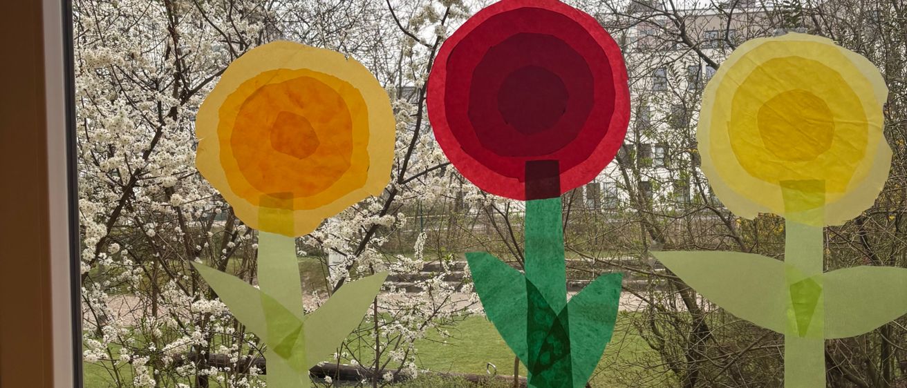 Drei Papierblumen, gelb, rot und gelb, sind an einem Fenster befestigt, mit kahlen Bäumen im Hintergrund.
