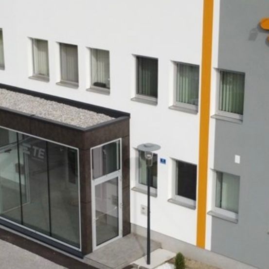 Ein Außenansicht eines modernen Gebäudes mit Glastüren und Fenstern. Das Gebäude hat ein graues und weißes Farbschema mit einem gelben Streifen an der Seite.