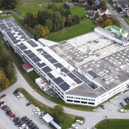 Luftaufnahme eines Industriegebäudes mit Solarmodulen auf dem Dach und einem davor liegenden vollen Parkplatz mit Autos.