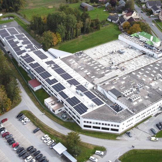 Luftaufnahme eines Industriegebäudes mit Solarmodulen auf dem Dach und einem davor liegenden vollen Parkplatz mit Autos.