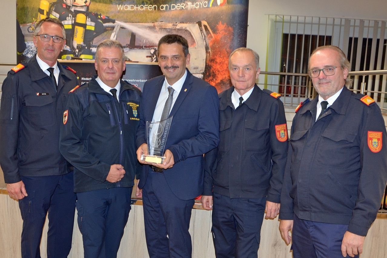 Fünf Männer in Uniformen stehen hinter einem Tresen, einer hält einen Preis. Dahinter ist ein Poster mit Feuerwehrleuten und einem Auto.