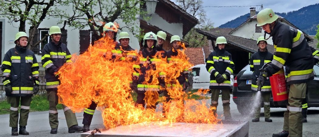 Eine Gruppe von Feuerwehrleuten trainiert mit einem kontrollierten Feuer, während ein Feuerwehrmann Wasser sprüht. Sie tragen Helme und Uniformen.