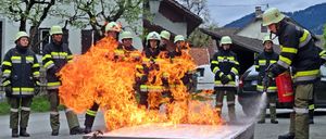 Eine Gruppe von Feuerwehrleuten trainiert mit einem kontrollierten Feuer, während ein Feuerwehrmann Wasser sprüht. Sie tragen Helme und Uniformen.