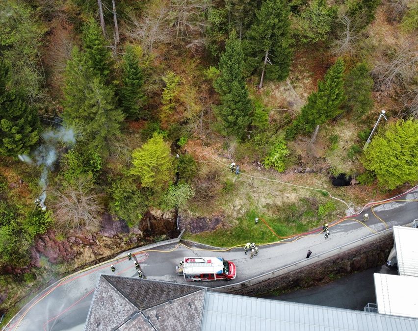 Luftaufnahme eines Waldes mit einer kurvenreichen Straße und Feuerwehrleuten, die einen Brand am Hang löschen.