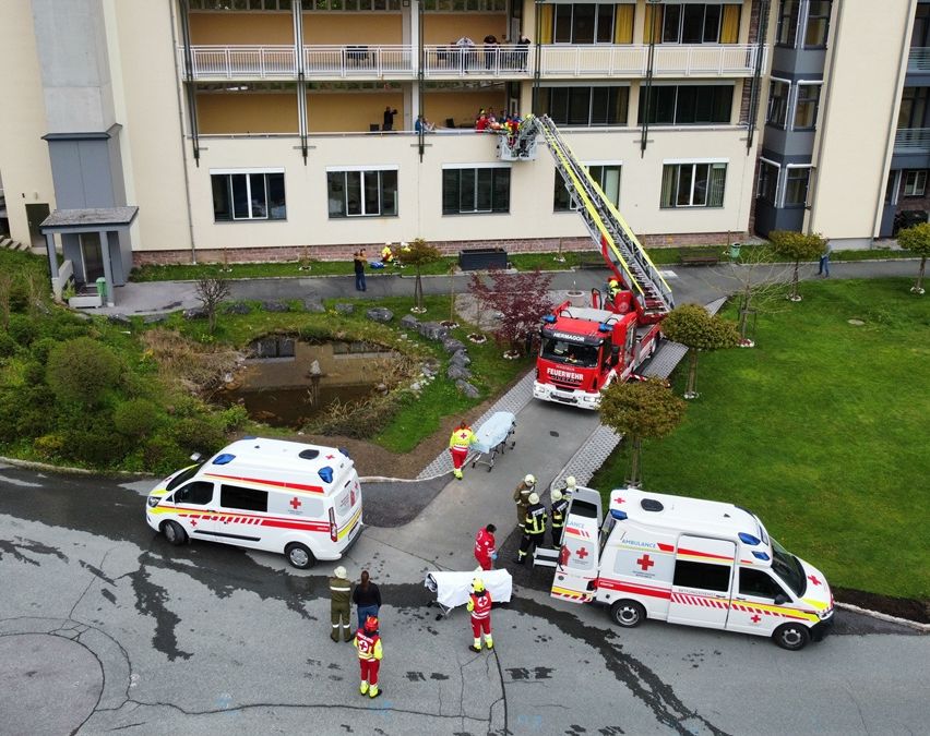 Eine Luftaufnahme einer Notfallszene mit zwei Rettungswagen, einem Feuerwehrwagen mit ausgefahrener Leiter und mehreren Rettungskräften um eine Person auf einer Trage.