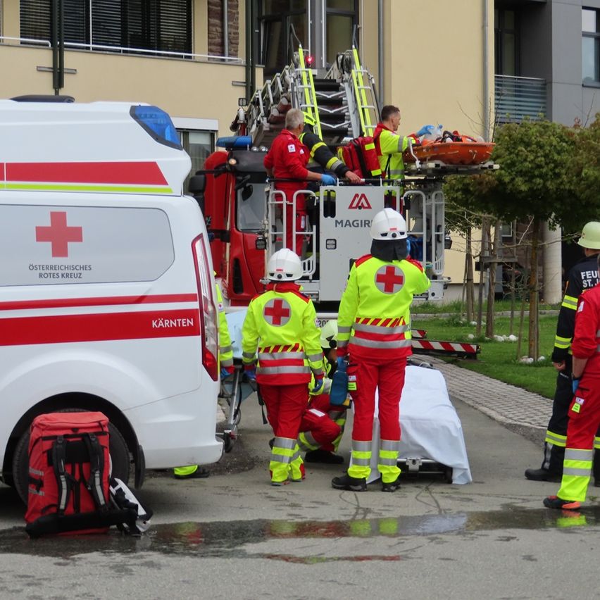 Rettungskräfte bringen einen Patienten von einem Feuerwehrwagen in einen Krankenwagen. Die Szene spielt sich vor einem Gebäude mit Treppen und Balkon ab.