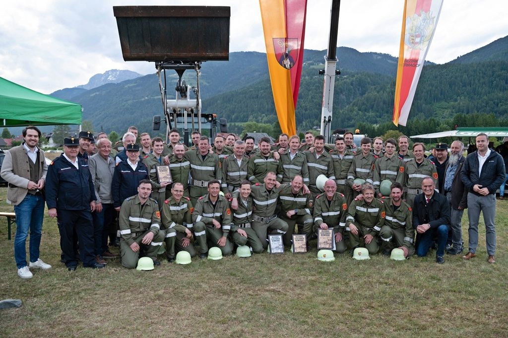 Eine Gruppe von Menschen in Militäruniformen und Helmen posiert für ein Foto auf einem Grasfeld. Dahinter befinden sich Berge und einige Fahrzeuge.